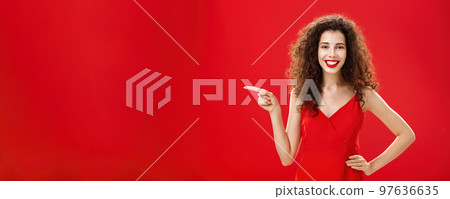 Girl suggesting ask her boyfriend. Charming elegant and stylish young female with curly hairstyle in red evening dress holding hand on hip and pointing left while showing friend where buy clothes 97636635