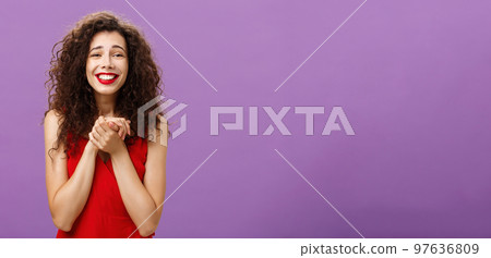 Woman happy receive award being touched and delighted holding palms together near breast smiling with tender and grateful expression standing with curly hairstyle in evening red dress over purple wall Woman happy receive award being touched and delighted holding palms together near breast smiling with tender and grateful expression standing with curly hairstyle in evening red dress over purple wall 97636809
