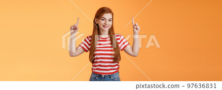 Cheerful beautiful redhead woman smiling invite try out new product, pointing up, show top copy space, grinning joyfully, rejoice introduce cool thing, stand orange background casual urban outfit 97636831
