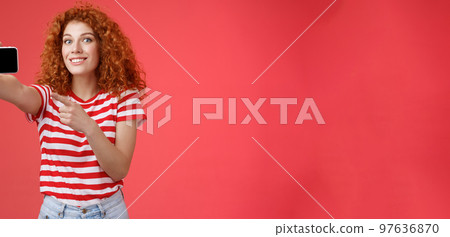 Excited happy good-looking redhead curly female show horizontal smartphone screen pointing display gadget smiling pleased proud beat friends score game standing red background 97636870