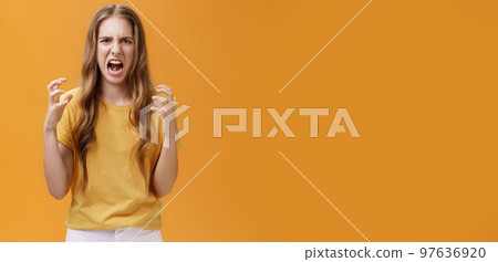 Portrait of irritated cute european female squeezing hands in fists with anger frowning and making hateful face standing annoyed and pissed over orange background with furious look 97636920