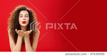 Tender and gentle stylish caucasian girl with curly hairstyle and red lipstick bending towards camera with slight smile closed eyes and palms near folded lips blowing kiss at camera romantically Tender and gentle stylish caucasian girl with curly hairstyle and red lipstick bending towards camera with slight smile closed eyes and palms near folded lips blowing kiss at camera romantically 97636999
