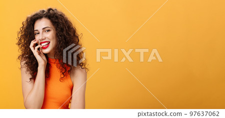 Waist-up shot of charming silly and carefree feminine woman with curly hairstyle in cropped top biting finger and smiling sensually and flirty at camera looking sexy and daring over orange background Waist-up shot of charming silly and carefree feminine woman with curly hairstyle in cropped top biting finger and smiling sensually and flirty at camera looking sexy and daring over orange background 97637062
