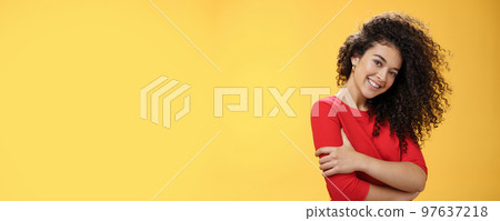 Waist-up shot of tender feminine and gentle woman with curly hairstyle combed to right side, tilting head and smiling flirty making romantic gazed at camera hugging herself over yellow background Waist-up shot of tender feminine and gentle woman with curly hairstyle combed to right side, tilting head and smiling flirty making romantic gazed at camera hugging herself over yellow background 97637218