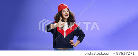 Nice work, keep up. Delighted and supportive charming sportswoman in winter beanie, sweatshirt and watch showing thumbs up with satisfied smile, giving approval, liking and cheering great effort 97637271