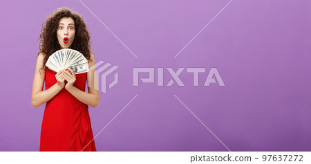 Studio shot of woman being lucky in casino winning lots of cash holding money in hands saying wow with folded lips staring impressed and excited over purple wall in evening red dress Studio shot of woman being lucky in casino winning lots of cash holding money in hands saying wow with folded lips staring impressed and excited over purple wall in evening red dress 97637272