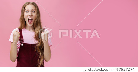 Indoor shot of slim funny and pretty young girl in corduroy overalls clenching fists in yes and triumph yelling from amazement and joy popping eyes being astonished by sudden win of favorite team Indoor shot of slim funny and pretty young girl in corduroy overalls clenching fists in yes and triumph yelling from amazement and joy popping eyes being astonished by sudden win of favorite team 97637274