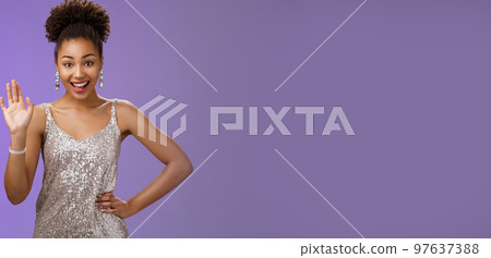 Friendly elegant african-american young 20s woman wearing elegant silver glittering dress hold hand waist waving hello greeting gesture say hi informally meet friend talking blue background Friendly elegant african-american young 20s woman wearing elegant silver glittering dress hold hand waist waving hello greeting gesture say hi informally meet friend talking blue background 97637388