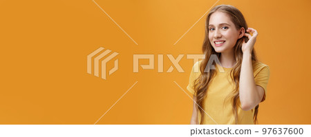 Cute and tender flirty young woman with cute wavy hairstyle flicking hair strand behind ear gazing at camera with timid sensual smile glancing with admiration at guy over orange background Cute and tender flirty young woman with cute wavy hairstyle flicking hair strand behind ear gazing at camera with timid sensual smile glancing with admiration at guy over orange background 97637600