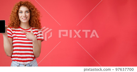 How about this app. Redhead curly friendly cute girl help friend find oufit online store hold smartphone pointing phone screen smiling broadly recommend social media photo filter, red background 97637700