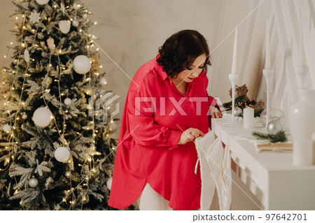 A woman in a red shirt in the room looks at a gift in a sock from Santa Claus, decorated for Christmas. Home New Year's atmosphere 97642701