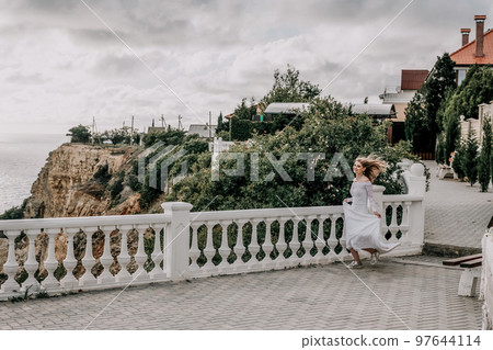 Romantic bride, blond girl in white wedding dress with open shoulders posing on open terrace with backdrop of the sea and rocks. Stylish young woman standing on terrace and looking on ocean at sunset 97644114