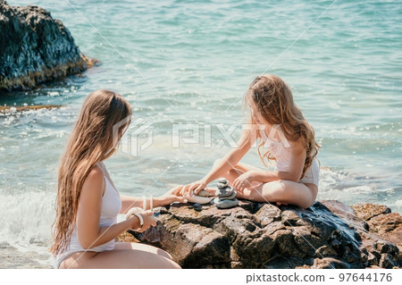 Woman with daughter bilds stones pyramid on seashore on a sunny day on the blue sea background. Happy family holidays. Pebble beach, calm sea. Concept of happy vacation on the sea, meditation, spa 97644176