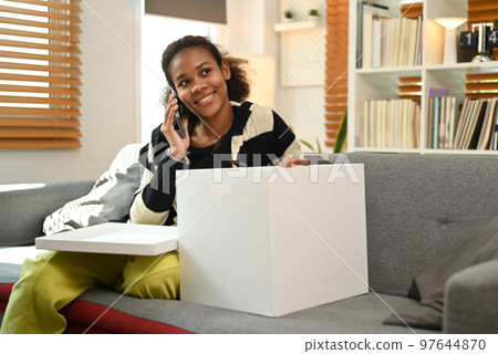 Smiling young woman talking on mobile phone and unpacking box on couch at home. Shopping online, good delivery concept 97644870
