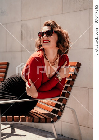 Portrait of a smiling woman on the street. An attractive woman with glasses, a red blouse and a black skirt is sitting on a bench on the street smiling. 97647565
