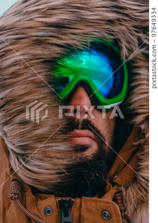 Headshot photo of a man in a cold snowy area wearing a thick brown winter jacket, snow goggles and gloves. Life in cold regions of the country. 97649334