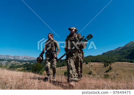 A sniper team squad of soldiers is going undercover. Sniper assistant and team leader walking and aiming in nature with yellow grass and blue sky. Tactical camouflage uniform. 97649475