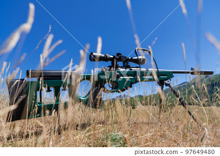 A green military sniper rifle with a scope for long distance tactical modern warfare in yellow grass blue sky 97649480