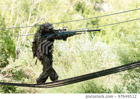 A military man or airsoft player in a camouflage suit sneaking the rope bridge and aims from a sniper rifle to the side or to target 97649512