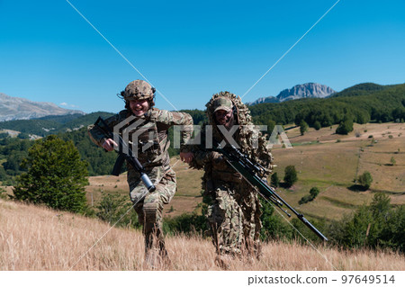 A sniper team squad of soldiers is going undercover. Sniper assistant and team leader walking and aiming in nature with yellow grass and blue sky. Tactical camouflage uniform. A sniper team squad of soldiers is going undercover. Sniper assistant and team leader walking and aiming in nature with yellow grass and blue sky. Tactical camouflage uniform. 97649514