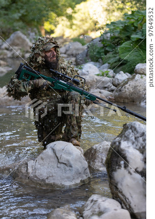 A military man or airsoft player in a camouflage suit sneaking the river and aims from a sniper rifle to the side or to target. A military man or airsoft player in a camouflage suit sneaking the river and aims from a sniper rifle to the side or to target. 97649532