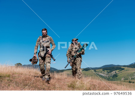 A sniper team squad of soldiers is going undercover. Sniper assistant and team leader walking and aiming in nature with yellow grass and blue sky. Tactical camouflage uniform. A sniper team squad of soldiers is going undercover. Sniper assistant and team leader walking and aiming in nature with yellow grass and blue sky. Tactical camouflage uniform. 97649536