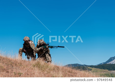 A sniper team squad of soldiers is going undercover. Sniper assistant and team leader walking and aiming in nature with yellow grass and blue sky. Tactical camouflage uniform. A sniper team squad of soldiers is going undercover. Sniper assistant and team leader walking and aiming in nature with yellow grass and blue sky. Tactical camouflage uniform. 97649548