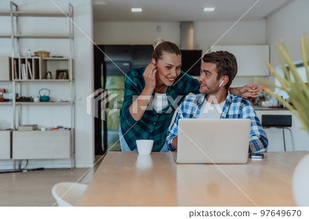 A young married couple is talking to parents, family and friends on a video call via a laptop while sitting in the living room of their modern house 97649670