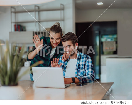 A young married couple is talking to parents, family and friends on a video call via a laptop while sitting in the living room of their modern house 97649673