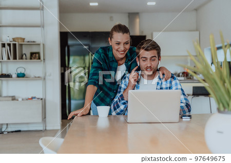A young married couple is talking to parents, family and friends on a video call via a laptop while sitting in the living room of their modern house 97649675