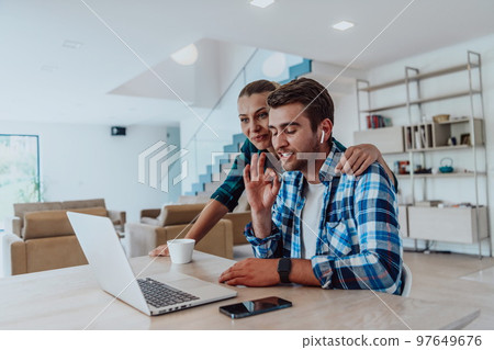 A young married couple is talking to parents, family and friends on a video call via a laptop while sitting in the living room of their modern house 97649676