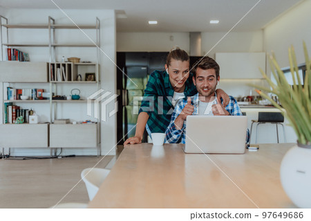A young married couple is talking to parents, family and friends on a video call via a laptop while sitting in the living room of their modern house 97649686