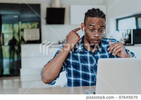 African American man in glasses sitting at a table in a modern living room, using a laptop for business video chat, conversation with friends and entertainment African American man in glasses sitting at a table in a modern living room, using a laptop for business video chat, conversation with friends and entertainment 97649870