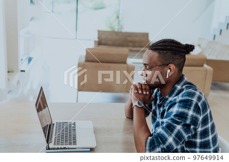 African American man in glasses sitting at a table in a modern living room, using a laptop for business video chat, conversation with friends and entertainment African American man in glasses sitting at a table in a modern living room, using a laptop for business video chat, conversation with friends and entertainment 97649914