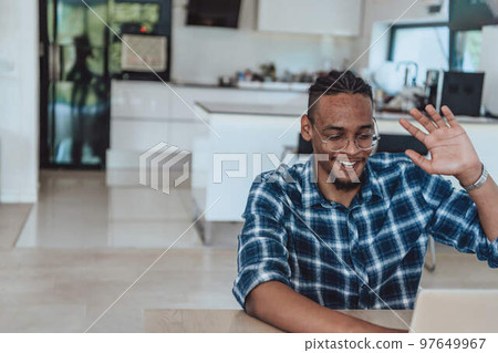 African American man in glasses sitting at a table in a modern living room, using a laptop for business video chat, conversation with friends and entertainment African American man in glasses sitting at a table in a modern living room, using a laptop for business video chat, conversation with friends and entertainment 97649967