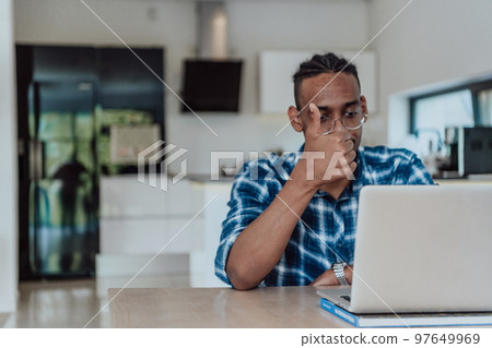African American man in glasses sitting at a table in a modern living room, using a laptop for business video chat, conversation with friends and entertainment African American man in glasses sitting at a table in a modern living room, using a laptop for business video chat, conversation with friends and entertainment 97649969