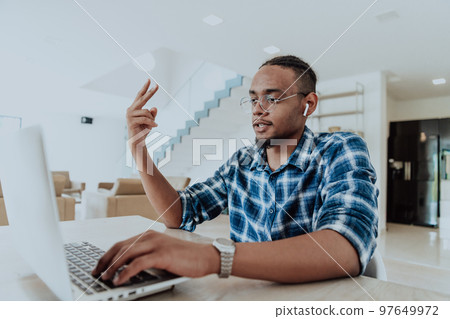 African American man in glasses sitting at a table in a modern living room, using a laptop for business video chat, conversation with friends and entertainment African American man in glasses sitting at a table in a modern living room, using a laptop for business video chat, conversation with friends and entertainment 97649972