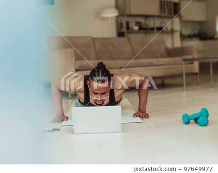 Young African American man working push-ups in the living room while watching online training on laptop Young African American man working push-ups in the living room while watching online training on laptop 97649977