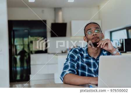 African American man in glasses sitting at a table in a modern living room, using a laptop for business video chat, conversation with friends and entertainment African American man in glasses sitting at a table in a modern living room, using a laptop for business video chat, conversation with friends and entertainment 97650002