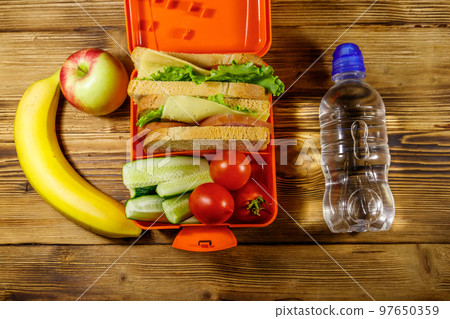 Bottle of water, apple, banana and lunch box with sandwiches and fresh vegetables on a wooden table. Top view Bottle of water, apple, banana and lunch box with sandwiches and fresh vegetables on a wooden table. Top view 97650359