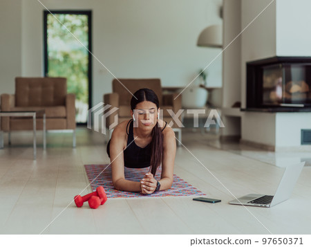 A young woman in sportswear working a plank in the living room while participating in online training via a laptop A young woman in sportswear working a plank in the living room while participating in online training via a laptop 97650371