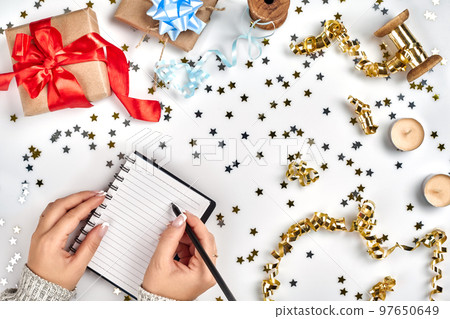 Festive decorations, delicate wavy ribbons, metallic star shaped confetti and notebook with wish list on white table, flat lay style. Christmas holidays decoration concept. 97650649
