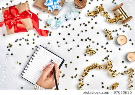 Festive decorations, delicate wavy ribbons, metallic star shaped confetti and notebook with wish list on white table, flat lay style. Christmas holidays decoration concept. Festive decorations, delicate wavy ribbons, metallic star shaped confetti and notebook with wish list on white table, flat lay style. Christmas holidays decoration concept. 97650650