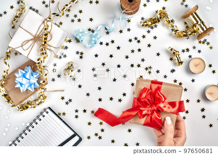 Female hands holding present with red bow on white background with delicate wavy ribbons and metallic star shaped confetti. Flat lay style 97650681