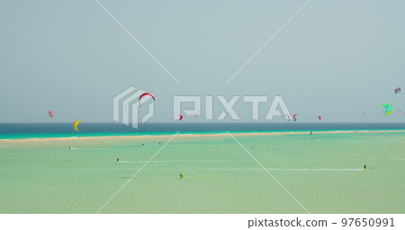 Kitesurfing on the tropical beach of Playa Del Mal Nombre, Fuerteventura, Canary Islands. People engaged in kiteboarding turquoise sea. The men on the board swims fast on the waves in shallow water. Kitesurfing on the tropical beach of Playa Del Mal Nombre, Fuerteventura, Canary Islands. People engaged in kiteboarding turquoise sea. The men on the board swims fast on the waves in shallow water. 97650991