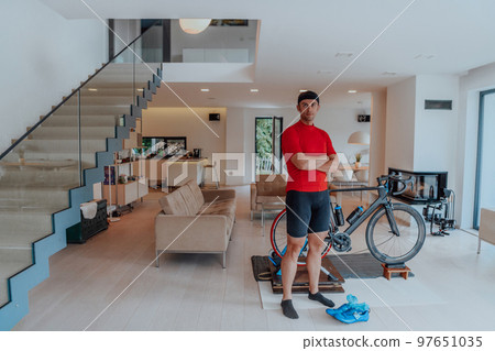 Photo of a determined triathlete standing in a modern large living room, behind him is his training bike 97651035