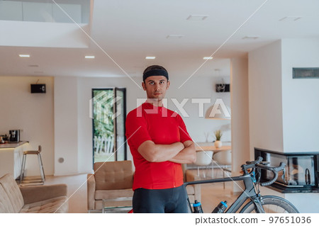 Photo of a determined triathlete standing in a modern large living room, behind him is his training bike Photo of a determined triathlete standing in a modern large living room, behind him is his training bike 97651036