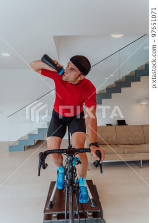 An athlete drinking water while riding a triathlon bike on a simulation machine in a modern living room. Training in pandemic conditions. An athlete drinking water while riding a triathlon bike on a simulation machine in a modern living room. Training in pandemic conditions. 97651071