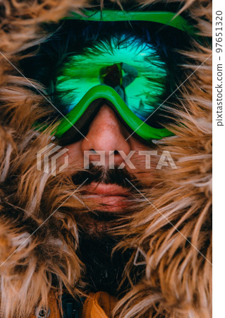 Headshot photo of a man in a cold snowy area wearing a thick brown winter jacket, snow goggles and gloves. Life in cold regions of the country. 97651320