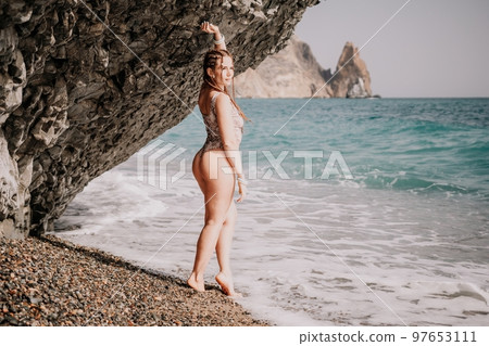 A tourist woman in bikini on beach enjoys the turquoise sea during her summer holiday. Beach vacation. Woman with braids dreadlocks standing with her arms raised near basalt rock enjoying beach ocean A tourist woman in bikini on beach enjoys the turquoise sea during her summer holiday. Beach vacation. Woman with braids dreadlocks standing with her arms raised near basalt rock enjoying beach ocean 97653111